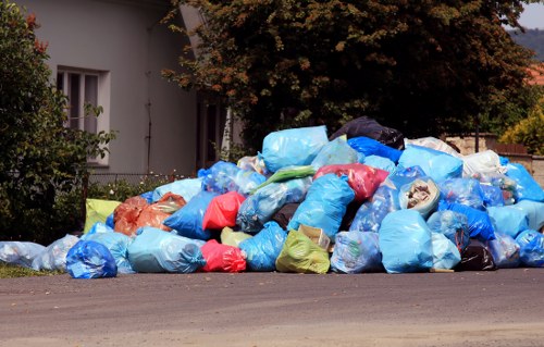 A large pile of tightly packed rubbish bags is situated on the edge of a paved street in a residential area, with some bags appearing full and slightly bulging. The bags are predominantly blue, with a few red, orange, green, and yellow bags interspersed, made of plastic material and showing signs of being used for waste collection. Behind the pile, there's a white residential building with vertical window bars and a well-maintained garden featuring shrubs and a tree with dark red and brown leaves. To the right, more foliage from a larger tree extends upward, partially obscuring the sky. The scene is lit by natural daylight, with soft shadows cast around the rubbish bags, indicating clear weather. This image illustrates typical rubbish accumulation in a suburban setting, relevant to local rubbish removal services like those offered by Garden Clearance Hillingdon, in the Hillingdon area. The overall environment suggests a quiet street near residential premises, highlighting the need for efficient garden clearance and rubbish disposal solutions.